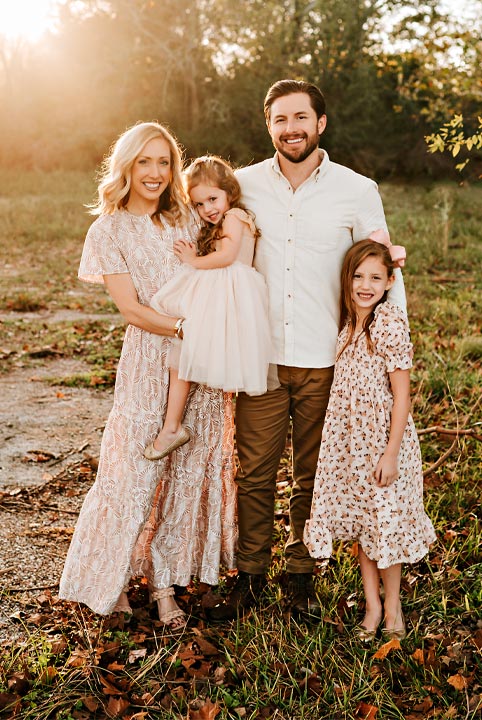 Family of four standing outdoors at sunset, smiling together in a natural, grassy setting. The parents stand side by side while the mother holds their younger daughter, and the older daughter stands beside them. Soft golden light creates a warm, joyful atmosphere.