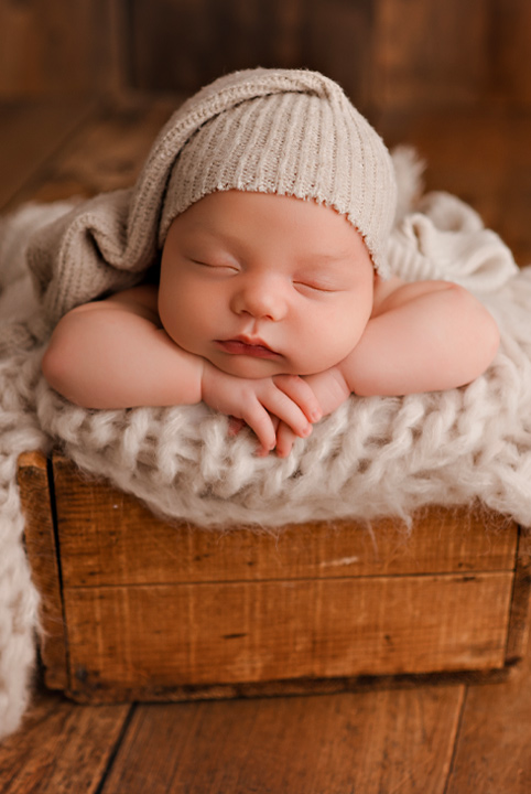 Newborn baby sleeps in wooden box lined with soft plush blankey. It's arms resting under its chin and a soft cap covers its head.